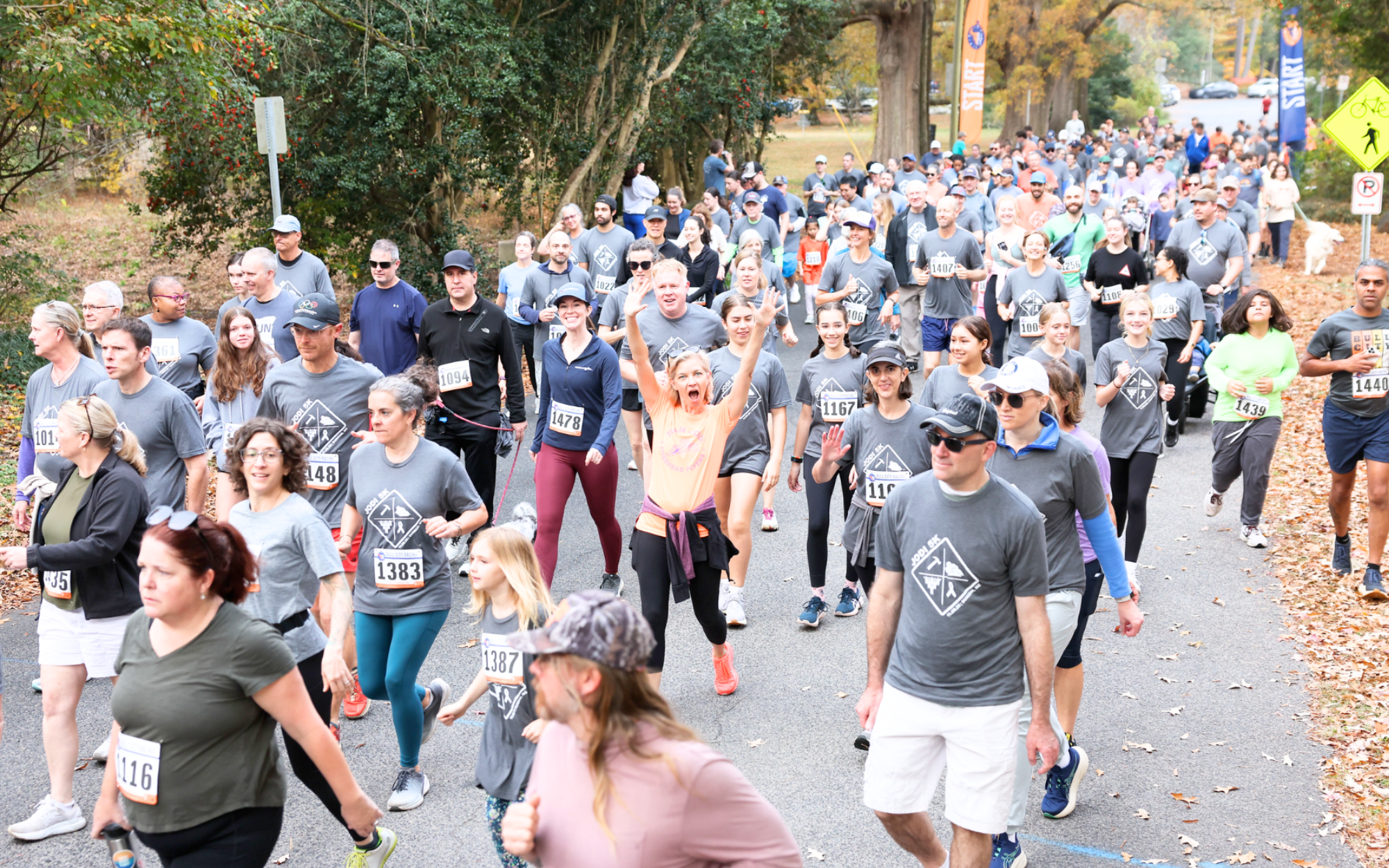 Hundreds of runners and walkers in gray Jodi 5K shirts heading out from the start line on a tree-lined road in the fall