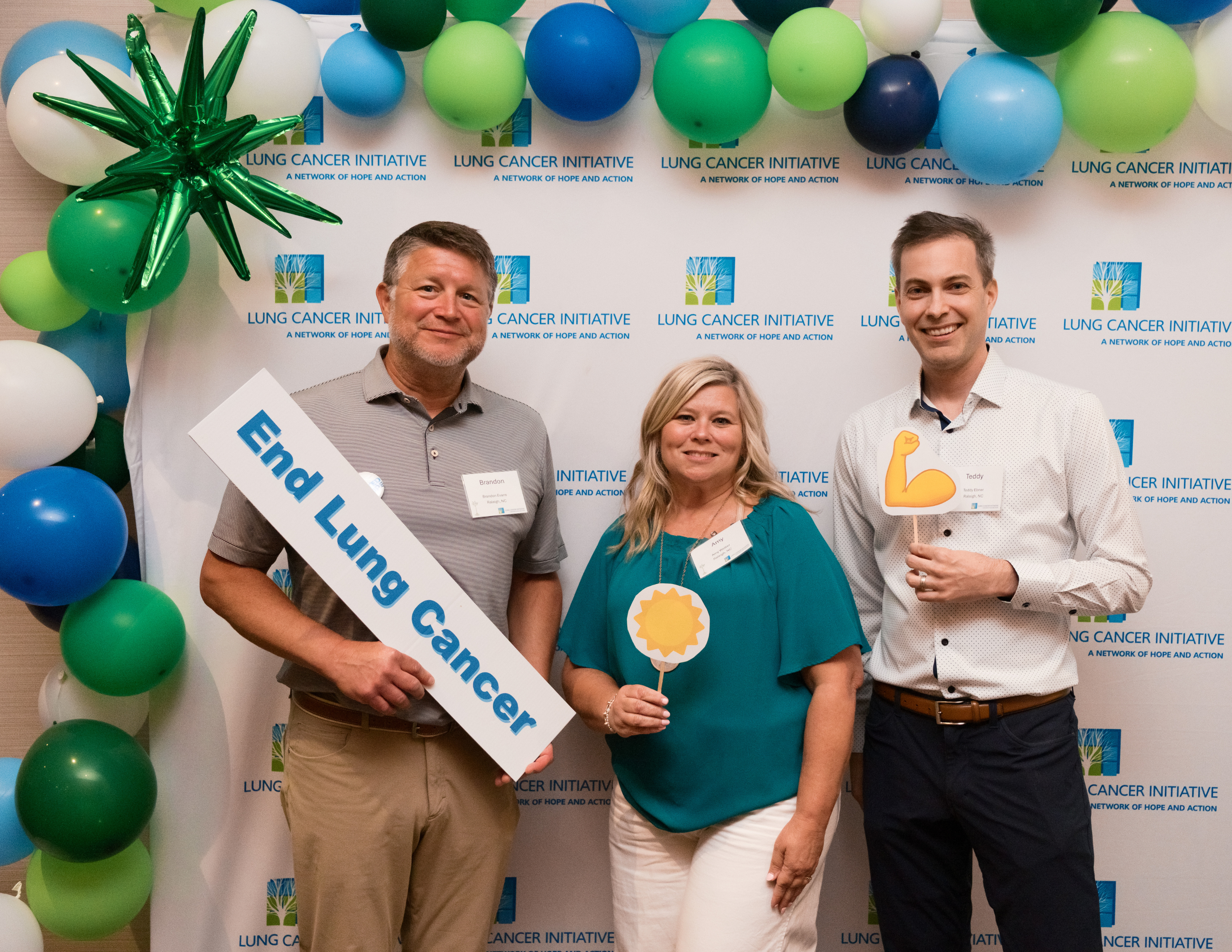 Three supporters smiling in front of a Lung Cancer Initiative backdrop, one holding an End Lung Cancer sign
